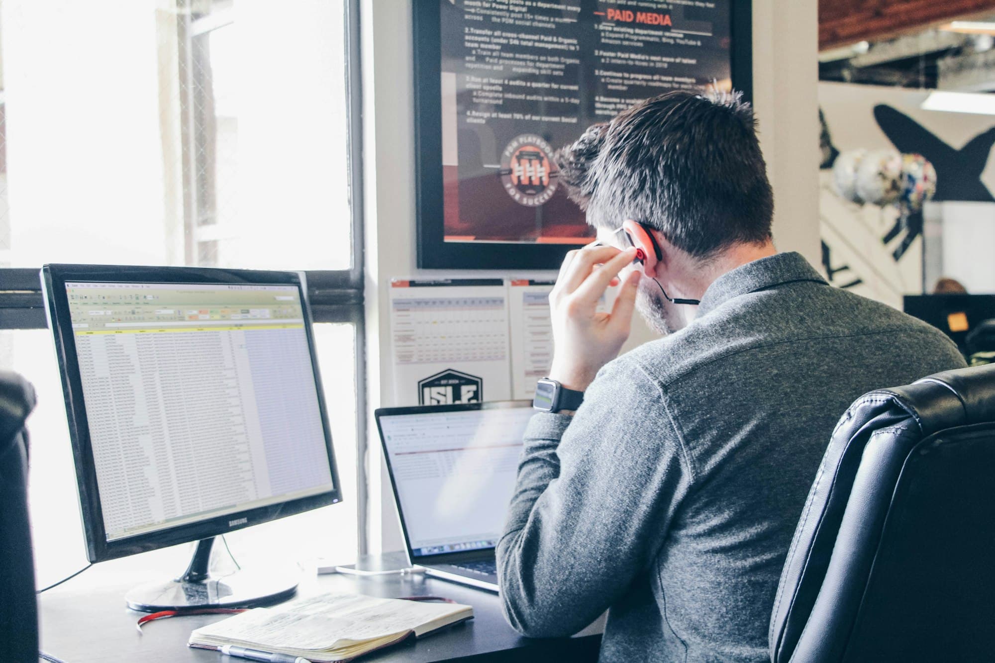 An accountant at his desk working through a client spreadsheet, with a laptop open beside the monitor